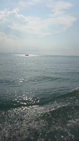 The video shows a view from a moving boat on the water. The camera is angled downwards towards the churning wake of the boat. In the distance, there are several other boats and a faint horizon line. The sky is partly cloudy with sunlight reflecting off the water. The overall impression is of a calm, sunny day at sea.