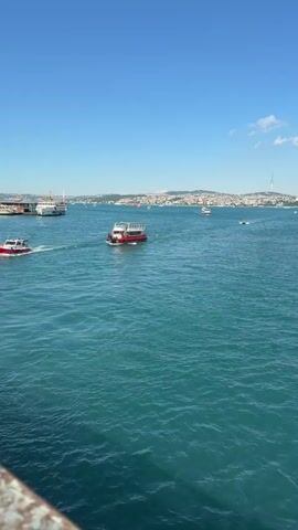 A video shows a body of water with several boats. In the foreground, the water is choppy with a slight ripple. A small red and white boat is moving from left to right in the middle ground, leaving a wake. Further in the background, a larger red and white ferryboat is also moving from left to right. A larger white ship is docked on the left side of the frame. The background shows a cityscape with buildings and a television tower on a hill. The sky is clear and blue with a few scattered clouds. The video is shot from a slightly elevated perspective.