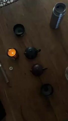The video shows a wooden table with several objects arranged on it. There is a lit candle in a striped glass holder, two small teapots (one dark grey and one reddish-brown), two small tea cups, a glass of tea, a stainless steel thermos, an incense stick, and a small white disc-shaped object. A lighter and a box of tea bags are visible in the upper left corner of the frame. The scene is dimly lit. The camera slowly zooms out.