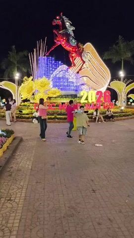 At night, a low-angle shot captures a bustling public space. A brightly lit display takes center stage, featuring a stylized horse figure and the year "2026". The display is adorned with flowers and other decorative elements. People of varying ages stroll around, some taking photos. A girl with braided hair stands in the foreground, gazing at the display. A boy runs toward the camera. The ground is paved with interlocking bricks. Palm trees punctuate the background, their silhouettes barely visible against the dark sky.