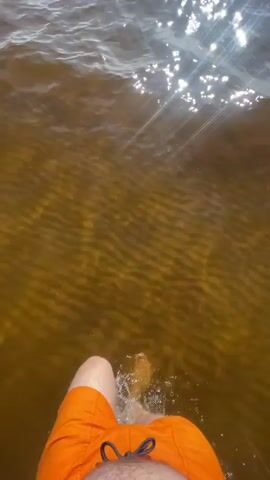 The video shows a person's legs as they walk into murky brown water. The sun reflects brightly off the surface of the water, creating a shimmering effect. The person is wearing orange swim trunks, and their bare feet are visible as they move through the water, creating ripples and splashes. The camera is positioned to look down at the person's legs and feet.