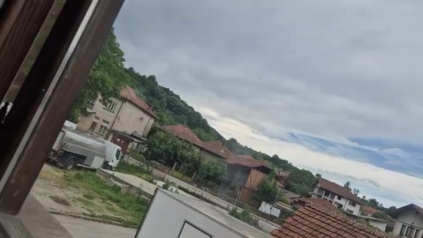 The video shows a tilted view of a rural village with houses and a hill in the background. A white truck with a large tank is parked in the foreground. The sky is cloudy with some patches of blue. There are no people or animals visible in the video. The overall scene suggests a quiet, possibly overcast day in a village setting.