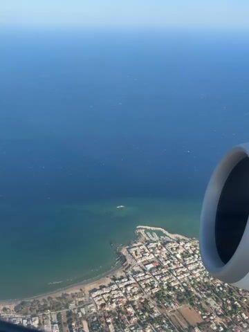 This is an aerial view from an airplane window looking down at a coastal town and the sea. Part of the airplane's engine is visible in the foreground on the right side of the frame. The town has white buildings and a harbor with boats. The sea is a deep blue and transitions to a greener shade closer to the shore. The water has a slightly rippled surface, indicating some movement or wind. The sky above is clear and bright blue.