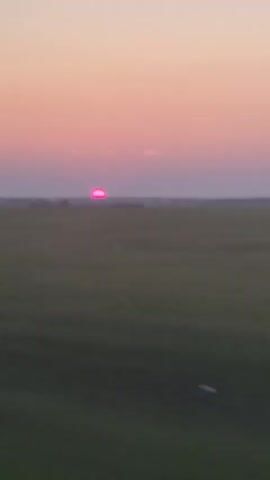 A time-lapse video shows a sunset over a field. The sun, which appears pink, is low on the horizon. The sky transitions from a pale pink to a deeper shade of purple as the sun dips below the horizon. The field below is a muted green and brown.