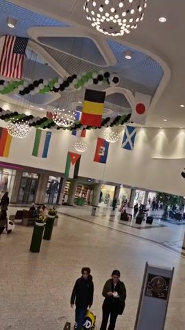 The video pans across a large, modern indoor space that appears to be a shopping mall or a public atrium. The ceiling is high and features architectural elements with integrated lighting. Several large, spherical light fixtures hang from the ceiling, emitting a bright, diffused light.  Decorations are visible throughout the space, including strings of balloons in green, black, and white, interspersed with what appear to be soccer balls. Several national flags are displayed: the United States, Belgium, Japan, Palestine, Mexico, and Scotland are clearly identifiable.  A U.S. flag hangs prominently in the upper left. The floor is tiled. In the mid-ground, there is a second level with a railing, where several people are seated at tables in what looks like a cafe or restaurant.  On the ground level, various shops are visible with displays and entrances. Several individuals are walking through the space. Two people are walking towards the camera in the foreground. In the lower right corner, a sign with a circular emblem is visible. The overall atmosphere suggests a public gathering or event, possibly related to a sporting competition given the flags and soccer ball decorations.