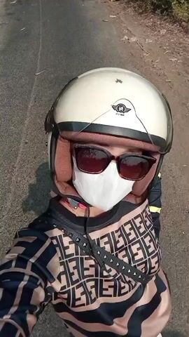 A first-person perspective video shows a person wearing a cream-colored helmet, sunglasses, and a white face mask. The person is also wearing a long-sleeved shirt with a black and beige repeating geometric pattern, adorned with a black strap with silver studs across the chest. They appear to be riding a motorcycle or scooter on an asphalt road with dry vegetation and trees on either side. The video captures the rider's view of the road ahead and the surrounding environment, with the sky visible through the branches of trees. The rider is speaking in Indonesian, saying "I'm walking with friends."