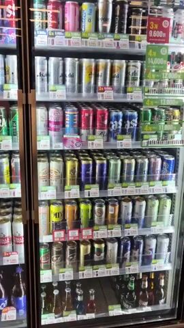 The video shows a tall refrigerator filled with cans and bottles of various beverages. The refrigerator has multiple shelves stocked with products that appear to be alcoholic drinks, such as beer and possibly soju. Some shelves have price tags displayed in Korean currency. The refrigerator door has a handle that is partially visible. The lighting suggests an indoor setting, likely a convenience store or supermarket. There are no people visible in the video.