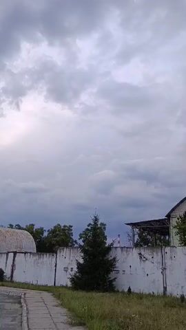 The video shows a wide shot of an outdoor industrial or abandoned site under a cloudy sky. In the foreground, a concrete sidewalk runs along the left side of the frame, bordered by dry grass and weeds. A white, weathered concrete wall extends across the middle ground, with various industrial structures visible behind it. To the left, a large, curved, metallic structure with a rough, textured surface is partially visible. A small, dark green evergreen tree stands in front of the wall. Further to the right, behind the wall, are trees and the side of a long, low building with a sloping roof. The building is painted in sections of white and light green, with a distinctive triangular window near the peak of the roof. To the far right, more trees and foliage are visible. In the distance, a tall, slender structure, possibly a smokestack or industrial chimney, with a red band near its top, can be seen above the trees and buildings. The overall atmosphere is somewhat somber due to the overcast sky and the weathered appearance of the structures. The camera is stationary.