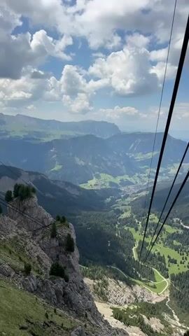 This is a first-person perspective video taken from a cable car ascending a mountain. The view looks down into a lush green valley dotted with trees and a winding road. In the distance, more mountains rise, some appearing hazy in the sunlight. The sky is blue with fluffy white clouds, suggesting fair weather. The time of day appears to be daytime, likely late morning or early afternoon. The atmosphere is serene and awe-inspiring, with the vastness of the landscape creating a sense of peace and wonder. The cable car itself is partially visible, with its thick black cables stretching diagonally across the frame. The focus is on the breathtaking natural scenery, highlighting the beauty of the mountainous region. The location is identified as Santa Cristina Gherdëina, Italy, known for its stunning Dolomite landscapes. There is no visible text in the video.