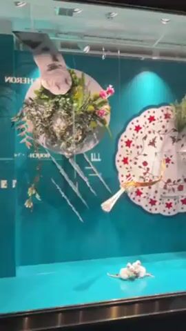 The video shows a window display with a blue backdrop and a collection of whimsical, gravity-defying objects.  On the left, a plate with a floral arrangement appears to float with silverware appearing to pierce the backdrop.  To the right, another ornate plate with red star patterns also floats, supporting a fabric drape and a glass. A teacup with a spoon extending from it hangs in mid-air.  At the base of the display, several small, white, plush mice huddle together. Red berries are scattered on the display surface. The lighting is bright and casts shadows on the display surface.