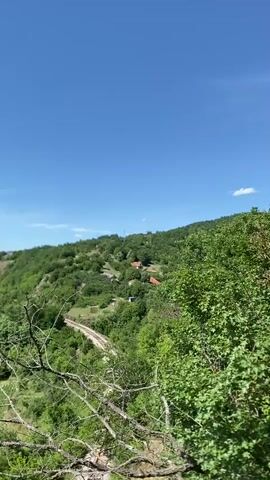 A drone shot pans over a lush green valley with a winding river and rolling hills under a bright blue sky with scattered clouds. In the foreground, dry tree branches frame the view. The landscape is dotted with trees, fields, and a few red-roofed houses. A railway track is visible winding through the valley. The sun is shining brightly, illuminating the vibrant green vegetation. The overall scene depicts a serene and picturesque natural environment.