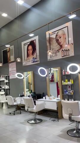 The video pans across the interior of a well-lit, modern hair salon. The walls are painted a dark gray, adorned with large posters featuring models with styled hair and product bottles. The floor is tiled in a light color. In the foreground, there are several styling stations, each with a white hydraulic chair, a mirror, and overhead ring lights. To the left, two white shampoo chairs with sinks are visible, suggesting a hair washing area. Behind the styling stations, shelves display various hair care products. A red reception desk with a textured front is situated in the center of the room, behind which a person wearing a black shirt and a red apron is partially visible. Further back, in the left section of the salon, a person is seated in a chair, appearing to be receiving a haircut. In the right section of the salon, there is a dark grey sectional sofa with an ottoman, and to the far left, a portion of a blue pool table is visible. The overall atmosphere is clean and professional, with multiple light sources creating a bright and inviting space.