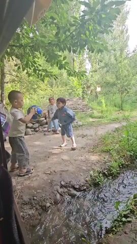 A group of three young children are playing outside near a stream. The child on the left is wearing a white t-shirt and grey pants and is holding a blue ball. The child in the middle is wearing a light blue t-shirt and pink pants. The child on the right is wearing a blue jacket and grey shorts. They are in a wooded area with trees and foliage. The ground is dirt and rocks. There is a stream of water flowing in the foreground. The children appear to be throwing the ball back and forth.