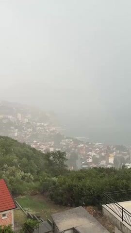 This is a still image captured from a video, showing a panoramic view of a coastal town during a heavy downpour. The scene is dominated by a dense, overcast sky, indicating rainy weather. The town, nestled along a hillside that slopes down towards the sea, is largely obscured by the mist and rain. Buildings, mostly of a white or light-colored facade with red-tiled roofs, are clustered together, creating a dense urban landscape.

The foreground features lush green vegetation, typical of a hillside environment, with trees and shrubs. In the lower section of the image, parts of residential structures are visible, including a red-tiled roof and concrete structures, suggesting a residential area overlooking the town and sea. The sea itself is a muted gray-green, blending into the hazy horizon.

The activity taking place is clearly a heavy rain shower. The presence of rain streaks in the video from which this still was taken would have conveyed the dynamic nature of the weather event. The overall mood evoked is one of a somber, wet day, with the mist and rain creating a sense of isolation and perhaps a bit of melancholy.

There is no visible text within the image. The location cues, such as the architecture and the coastal setting on a hilly terrain, align with the provided context of Sušćepan, Montenegro. The time of day is indeterminate due to the overcast conditions, but it appears to be daylight.