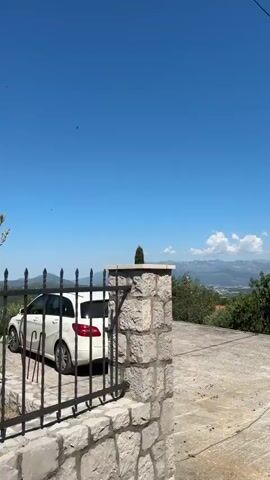 The video pans across a landscape under a clear blue sky. In the foreground, a white car is parked on a concrete surface. Behind the car, there is a stretch of trees and vegetation. Further in the distance, mountains are visible. The camera then moves to show a tall, slender tree, followed by more greenery and a glimpse of a building with a red roof. The video concludes by showing a metal fence bordering the property, with a concrete pillar.