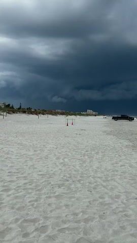 The video shows a wide shot of a beach with dark, stormy clouds overhead. The sand is light in color and shows tire tracks, indicating vehicles have been on the beach. Several vehicles, including a black pickup truck, are parked or driving on the sand near the water's edge. In the distance, buildings can be seen along the coastline. A few beach chairs are visible on the sand. The stormy clouds create a dramatic and ominous atmosphere. The video pans slightly to the right, showing more of the beach and the approaching storm.
