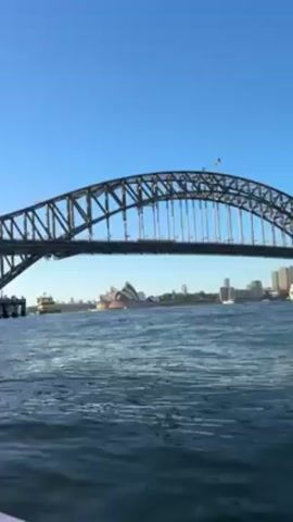 The video shows a view from a boat on the water, looking towards the Sydney Harbour Bridge and the Sydney Opera House on a clear, sunny day. The water is blue and has small waves. The bridge is a large steel arch structure, and the Opera House has its distinctive white, sail-like roofs. In the background, there is a cityscape with various buildings.
