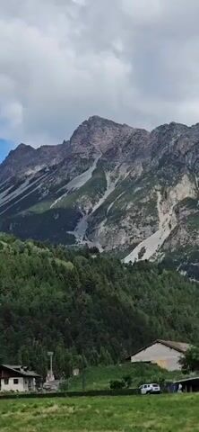 The video shows a panoramic view of a mountainous region with green forests and some houses. The mountains are tall and rugged, with some areas of bare rock and scree slopes. The sky is cloudy, but there are some patches of blue visible. The video pans across the landscape, offering different perspectives of the mountains and the village nestled below. In the foreground, there are some trees and a few parked cars. The overall impression is one of natural beauty and tranquility.