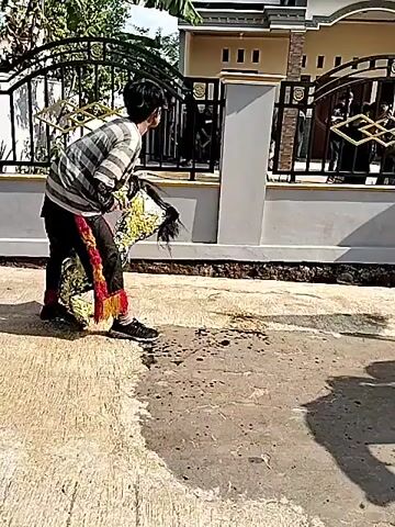 The video shows an outdoor scene with a group of people gathered near a house. The ground is wet and appears to have puddles and water stains. A person on the left is wearing a striped shirt, black pants, and red and yellow leg coverings. They are holding a basket-like object. Several other people are standing and interacting. One individual is holding a flag, which is red and white. There are trees and vegetation in the background, and the sky is visible. Some people are wearing casual clothing, while others appear to be in traditional attire. The atmosphere seems to be one of a local gathering or event.