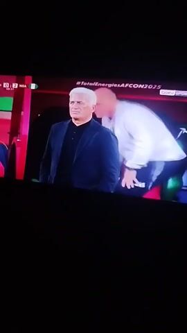 The video shows a recording of a TV screen broadcasting a soccer game. In the first shot, two men are seen. The man on the left has short white hair, a black shirt and a navy jacket. The man on the right is bald, wearing a white shirt and adjusting something with his hands. In the next shot, there is a man in the foreground wrapped in an Algerian flag, and wearing what appears to be a white jersey, sitting in a stadium seat. Behind him, there are other people wearing jerseys and a maroon shirt. There are logos visible on the apparel. The TV screen displays the game score.