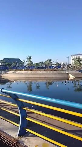 A vertical, slightly shaky video displays a calm body of water, reflecting the clear blue sky and a few scattered trees. In the background, a multi-story white building stands out, surrounded by smaller structures and more trees. The foreground shows a railing with blue and yellow horizontal bars, casting shadows on what appears to be a walkway. The scene suggests a public space, possibly a park or waterfront area, under bright daylight conditions. The camera pans slowly to the right, revealing more of the water and the distant shoreline.