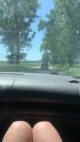 The video begins from the perspective of someone inside a car, looking out the windshield. The dashboard is visible in the foreground. The car appears to be driving on a rural road lined with trees. In the distance, another dark-colored car is visible ahead on the road. The sky is a clear, bright blue, suggesting it is daytime and likely sunny. Green fields and vegetation are visible on either side of the road, with lush trees providing shade along the roadside. There is a hint of what appears to be crops in the background fields.

The camera then pans to the right, showing more of the roadside scenery. A wooden pole with wires is visible, possibly an electrical or telephone pole, with a long wire hanging loosely. The vegetation on the right side of the road is dense and green. The scene is tranquil and appears to be a leisurely drive through the countryside on a pleasant day.

There is a sticker visible on the windshield that reads "GO SHOCK". Another small, rectangular black object is mounted on the dashboard.

The location context provided is Brusnytsia, Ukraine. The overall impression is that of a summer drive through a picturesque rural area of Ukraine.