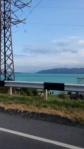 The video shows a view from a moving vehicle traveling along a road. On the left side of the road, there is a steep embankment with grass and some bushes. A road barrier with black and white stripes is visible along the embankment. A large electricity pylon stands prominently in the foreground. Beyond the embankment, there is a body of water that appears turquoise in color, with distant hills or mountains visible on the other side. The sky is blue with some scattered clouds. The vegetation includes trees and tall grasses. The overall scene suggests a scenic route, possibly near a lake or sea.