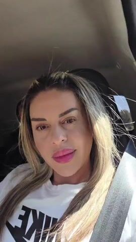 A woman with blonde hair and full lips wearing a white t-shirt with a Nike logo is in the front passenger seat of a car. She is looking at the camera. The background shows the interior of the car and a person in the back seat. The video is shot from a selfie perspective.