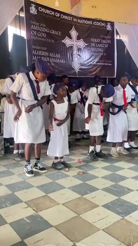The video shows a group of children, all dressed in matching white uniforms with blue caps and red accents, performing a synchronized dance in a church. They move in unison, following the rhythm of the music. The children are standing on a black and white checkered floor, and in front of a banner that displays the name of the church, "CHURCH OF CHRIST IN NATIONS (COCIN)". The banner also has text with "THEME 2026 AMAZING GRACE OF GOD (Ephesians 2:8-9)" and "ALHERIN ALLAH MAI BAN MAMAKI (Afisawa 2:8-9)". The camera angle is slightly high, giving a clear view of the dancers. The setting seems to be inside a church hall with white curtains. The children appear to be enjoying the performance.