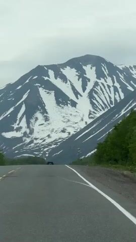 A car drives on a paved road towards a snow-capped mountain under a cloudy sky. Trees line the sides of the road.