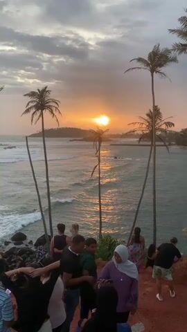 The video shows a group of people gathered on a clifftop overlooking the ocean at sunset. The sun is setting on the horizon, casting a warm glow over the water and the sky, which is filled with dramatic clouds. Palm trees are visible on the clifftop, and the ocean has gentle waves. Several people are taking photos or videos of the sunset, with some posing for selfies. One woman in a purple hijab is holding up her phone to take a picture. Another person is taking a photo of her. A few individuals are sitting on the edge of the cliff, looking out at the sea. The overall atmosphere is one of enjoyment and appreciation of the scenic view.