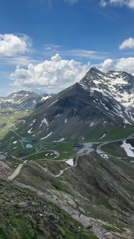 The video captures a panoramic view of a winding mountain road through a landscape of green hills and snow-capped peaks under a partly cloudy blue sky. The road is paved and features numerous switchbacks, indicating a significant elevation change. Several small lakes or ponds are visible nestled within the valleys. There are some buildings or structures visible near the road, possibly for tourism or services. The terrain is rugged, with rocky outcrops and patches of snow on the higher slopes. The overall impression is of a scenic, mountainous region, likely a popular tourist destination for its dramatic views and road access. There are no people or animals clearly visible, but a few vehicles are spotted on the road, suggesting it is open for travel.