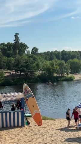 The video shows a sunny day at a lake with a sandy beach. In the foreground, a stand-up paddleboard leans against a blue pole. Several people are visible on the beach, some standing and some sitting. Three beach umbrellas, two with blue and white stripes and one with yellow and white stripes, are set up. A baby stroller is also present. Further back, on the lake, numerous individuals are engaged in stand-up paddleboarding. The lake is surrounded by lush green trees and a bright blue sky with scattered white clouds.