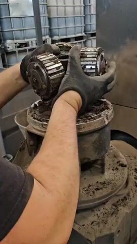 A person wearing black work gloves is holding a gear mechanism with two interlocking gears, covered in dirt, over an industrial machine that is also covered in dirt. The person removes the gear mechanism, revealing a splined shaft sticking out of the machine. The background shows a wall of large white industrial containers.