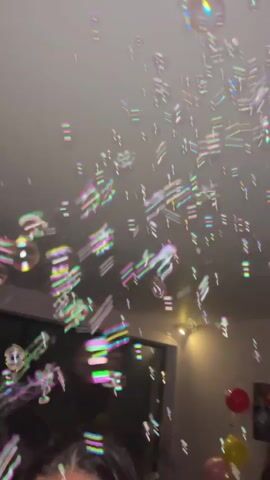 The video shows a room filled with many bubbles. Several balloons are visible in the background. A person can be seen trying to catch the bubbles with their hands.
