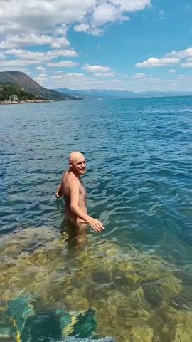 A bald man enters a body of water and swims. The man is in a natural state. The water is clear and the bottom is visible with rocks and seaweed. The man swims away from the camera. The sky is blue with white clouds. There are mountains in the background.