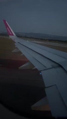 The video shows the wing of a Wizz Air airplane during takeoff or landing at night. The scene is an airport runway, with lights visible in the distance. The sky is dark, indicating it is either early morning or evening. The wing of the plane takes up most of the frame, with the "wizzair.com" logo visible on the tail fin. The plane appears to be moving, as suggested by the blurred lights and the dynamic perspective. The weather seems clear, with no visible rain or fog. The overall mood is one of travel and transition.