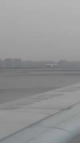 The video shows a plane on a runway, viewed from the wing of another plane. The weather appears to be overcast, with a gray sky and low visibility. In the background, a city skyline is visible. The plane on the runway is moving, but it is difficult to discern its exact direction.
