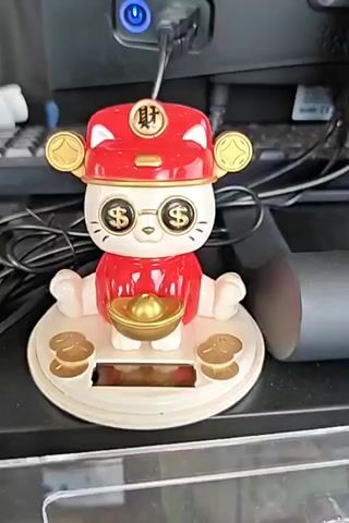 A close-up shot focuses on a solar-powered waving cat figurine with a red hat and outfit, gold dollar sign glasses, and holding a gold ingot. The figurine sits on a circular base with smaller gold ingots around it. The base has a solar panel visible at the bottom. The background is blurred, but appears to be a desk or workspace with computer equipment and cables. The figurine's right paw is waving.