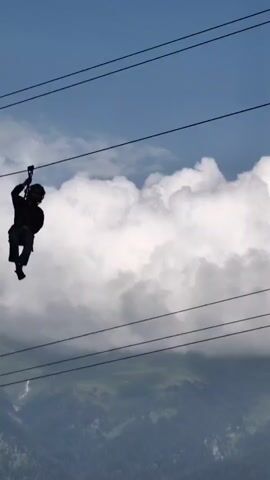 The video shows a person zip-lining across a mountainous landscape. The person is silhouetted against a bright sky with clouds. They are wearing a dark jacket, blue jeans, and sandals. The zip line is supported by cables stretched between unseen points. The background is a blur of green trees and mountains, suggesting movement and speed. The camera appears to be mounted on the zip line itself or is following the person closely.