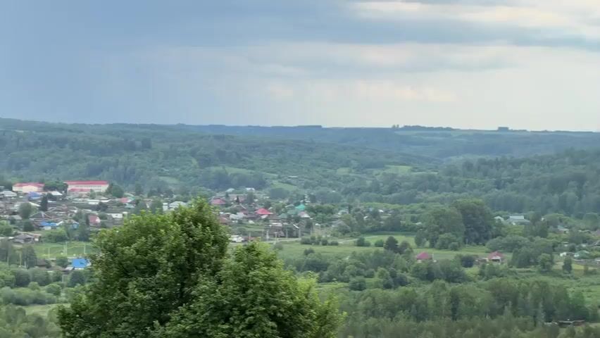 An aerial wide shot pans across a lush, green landscape. The foreground is dominated by the tops of dense trees. The mid-ground features rolling hills covered in forests, interspersed with a small village of houses with red and blue roofs. A narrow, winding river is visible. Further into the distance, more rolling hills are covered in forests, with some agricultural fields visible on the horizon. The sky is overcast with light clouds.