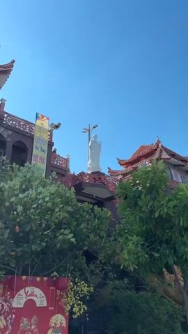 The video shows a bright blue sky with a large white statue of a person standing atop a building with red roofs. The statue is a person with their arms extended slightly outwards. The building appears to be a temple or shrine with ornate architectural features. In the foreground, there are green trees and foliage, obscuring some of the lower parts of the structure. A banner with text in a foreign language and some decorative elements hangs on the left side of the building. The scene appears to be outdoors, possibly on a sunny day.