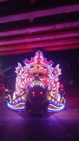 A nighttime video shows a large, elaborately decorated float shaped like a dragon moving under a bridge. The float is illuminated with colorful lights, primarily pink, purple, and blue. The dragon's head is at the front, with glowing red eyes and sharp teeth. The body of the float is adorned with ornate, flame-like structures and intricate patterns, also lit with LEDs. The bridge above is also illuminated with pink lights. The scene appears to be a parade or procession.