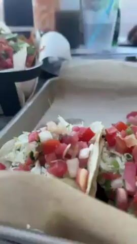 The video shows a table filled with food, including a basket of nachos topped with sour cream, guacamole, salsa, and jalapeños, and tacos filled with lettuce and chopped tomatoes. There are also bottles of hot sauce and a glass of water. The table is outdoors, with a view of a beach with several people on it. There are palm trees and light posts. People are walking along the boardwalk next to the beach.