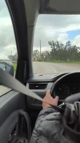 The camera, positioned from the driver's seat of a car, captures a first-person perspective of driving on a road. The driver's right hand is visible on the steering wheel, while their body is covered by a puffy black jacket. The seatbelt crosses the driver's chest. The dashboard and interior of the car are gray. Outside, the road is paved, and power lines run parallel to it. Trees are visible in the background against a cloudy, overcast sky. The car's speedometer is registering, indicating the vehicle is in motion. The overall setting suggests a rural or suburban environment.