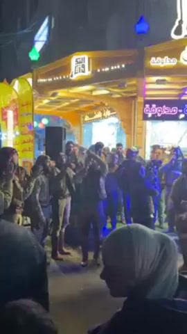 The video shows a crowd of people gathered on a street at night.  Most of the people appear to be men and several are wearing military-style clothing. They are clapping and chanting, with some raising their hands in the air. A person in the foreground is waving a Syrian flag. Buildings behind the crowd are decorated with bright, colorful lights and signage written in Arabic. The lighting on the street has a purplish hue. Several people are recording the scene with their phones.