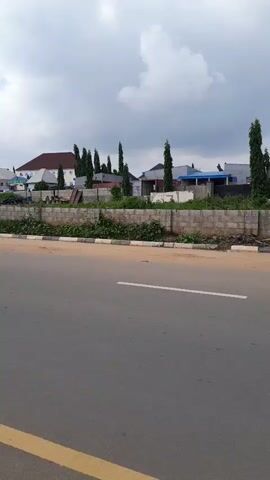 The video pans from right to left along an asphalt road. The road has dashed yellow lines marking the lanes and a solid yellow line on the far left side. On the right side of the road, there is a low stone wall, and beyond that, several modern houses and tall, slender trees. The sky is overcast with clouds. In the distance, a mountain range is visible. The overall scene appears to be a suburban or semi-rural area on a cloudy day.