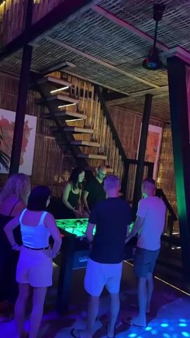 A group of six people are gathered around a foosball table in a dimly lit room. The room has a rustic ambiance with bamboo walls and a thatched ceiling. A staircase with illuminated steps is visible in the background. The lighting in the room is a mix of purple and green hues, and there are also blue disco lights projected onto the floor. Two men are actively playing foosball, while the other four people watch. One woman, wearing a white tank top and shorts, stands to the left, observing the game. Another woman, in a black dress, is also watching. A man in a black t-shirt stands behind the foosball table. Another man, in a grey t-shirt, is watching the game with his hands on his hips. The overall atmosphere appears to be social and recreational.