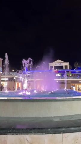 A nighttime shot focuses on a large, multi-tiered fountain with water jets shooting upwards. The fountain is illuminated with changing colored lights, cycling through blue, pink, purple, and green. In the background, a building with shops and restaurants is visible, with illuminated signs, including one that reads "SAFARI". People can be seen walking in front of the shops. The sky is dark, indicating it is night.