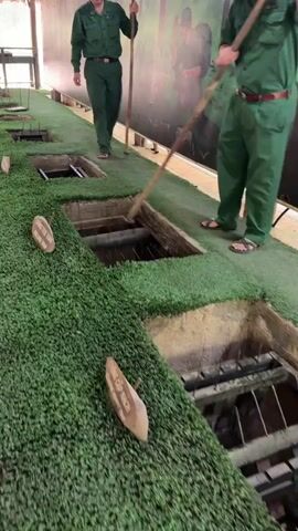 Two men in green military-style uniforms and sandals are demonstrating booby traps at what appears to be a historical exhibit, likely related to the Cu Chi Tunnels in Ho Chi Minh City, Vietnam. They are standing on artificial grass, next to several square holes in the ground.

One man, wearing a green hat, actively uses a long wooden pole to manipulate a mechanism within one of the holes, revealing a set of spikes. Another man, similarly dressed, stands nearby holding a pole. The holes are lined with wooden frames and contain various spike arrangements.

A small wooden sign in the foreground is visible, clearly inscribed with "CHONG CAP NAP" and "ROLLING TRAP", indicating the type of trap being shown. A partial view of another sign also starting with "CHONG" is seen. In the background, a mural depicting soldiers in a jungle setting is visible. The men appear to be guides or presenters, showcasing the historical defense mechanisms.